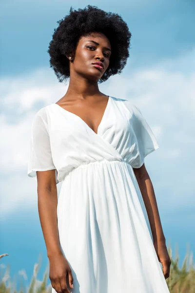 Vue à angle bas de la femme afro-américaine bouclée en robe blanche contre le ciel bleu — Photo de stock