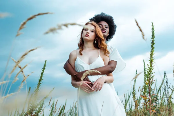 Enfoque selectivo de la mujer afroamericana abrazando chica pelirroja contra el cielo azul - foto de stock