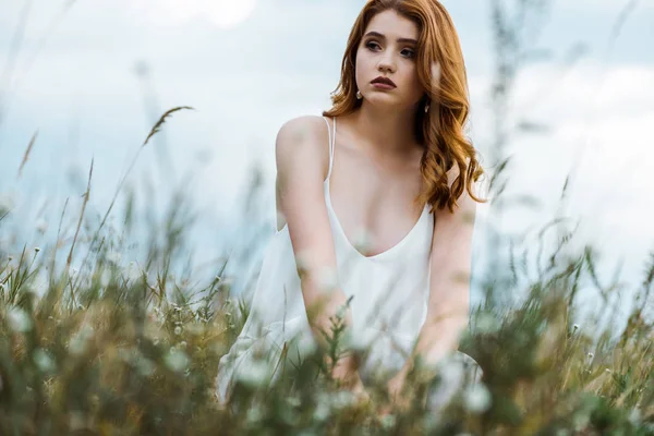Selective focus of young redhead woman in white dress sitting in grassy field — Stock Photo