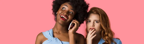 Foto panorámica de la mujer afroamericana feliz hablando en el teléfono inteligente cerca de chica pelirroja preocupado aislado en rosa - foto de stock