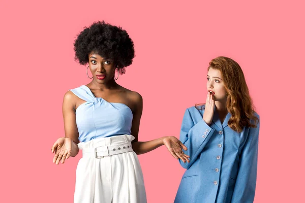 Überrascht rothaarige Mädchen mit Blick auf Afroamerikanerin zeigt Achselzucken Geste isoliert auf rosa — Stockfoto