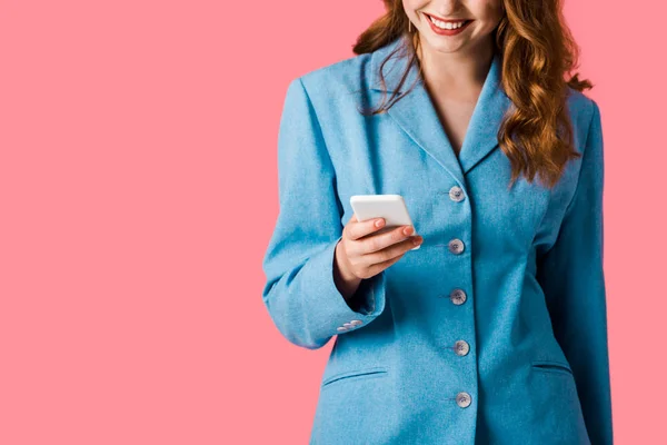 Vista recortada de chica pelirroja feliz usando teléfono inteligente aislado en rosa - foto de stock