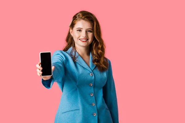 Chica pelirroja feliz sosteniendo teléfono inteligente con pantalla en blanco aislado en rosa - foto de stock