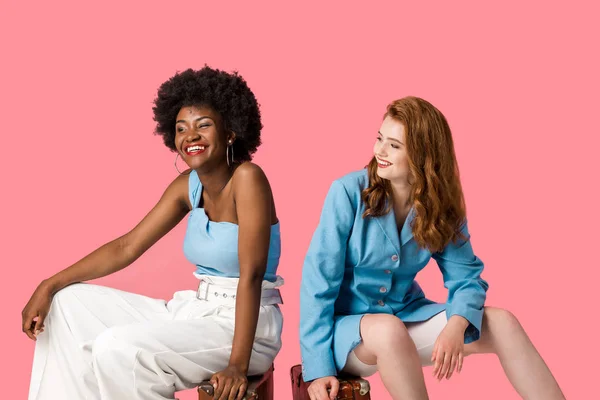 Happy multicultural women sitting on suitcases isolated on pink — Stock Photo