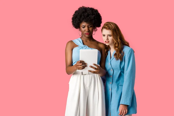 Displeased multicultural girls looking at digital tablet isolated on pink — Stock Photo