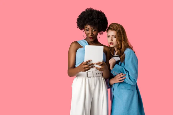 Pretty redhead girl holding credit card near african american woman with digital tablet isolated on pink — Stock Photo