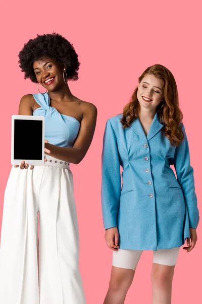 Menina ruiva alegre olhando para mulher afro-americana segurando tablet digital com tela em branco isolado em rosa — Fotografia de Stock