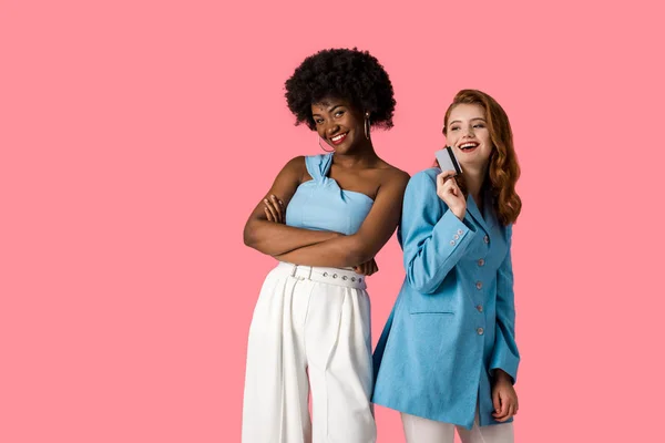 Happy redhead girl holding credit card near african american woman standing with crossed arms isolated on pink — Stock Photo