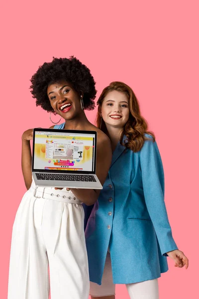 KYIV, UKRAINE - AUGUST 9, 2019: cheerful multicultural girls smiling near laptop with aliexpress website on screen isolated on pink — Stock Photo