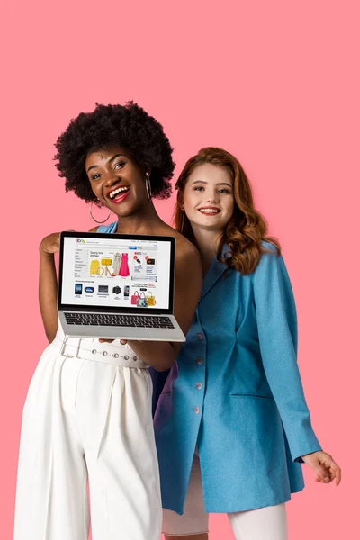 KYIV, UCRANIA - 9 de agosto de 2019: alegres chicas multiculturales sonriendo cerca de la computadora portátil con el sitio web ebay en la pantalla aislada en rosa - foto de stock