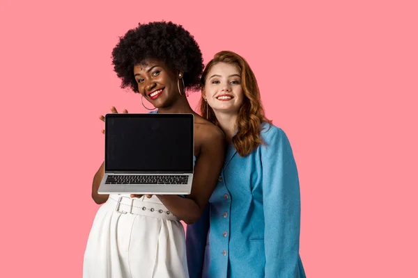 Meninas multiculturais alegres sorrindo perto do laptop com tela em branco isolada no rosa — Fotografia de Stock