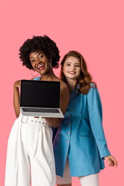 Heureuses filles multiculturelles souriant près d'un ordinateur portable avec écran vide isolé sur rose — Photo de stock