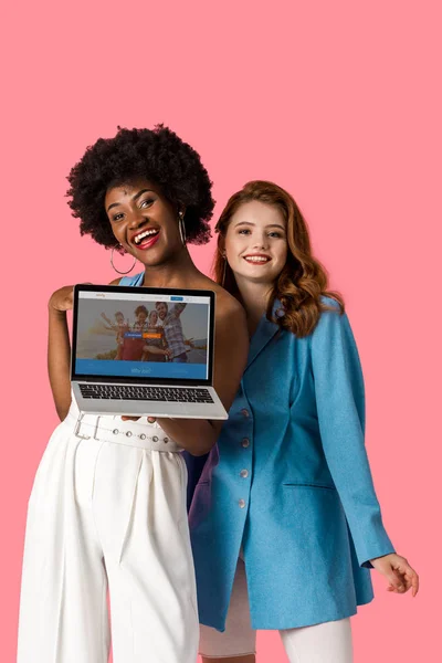 KYIV, UKRAINE - AUGUST 9, 2019: cheerful multicultural girls smiling near laptop with couchsurfing website on screen isolated on pink — Stock Photo