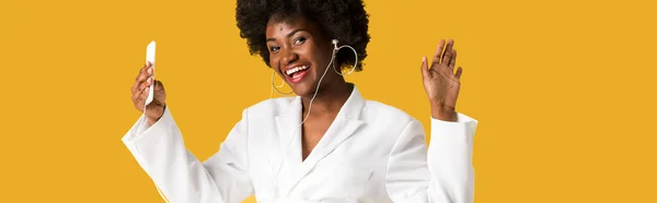 Panoramic shot of cheerful african american girl listening music while holding smartphone and gesturing isolated on orange — Stock Photo