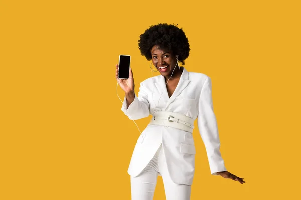 Sorridente menina afro-americana ouvir música enquanto segurando smartphone com tela em branco isolado na laranja — Fotografia de Stock