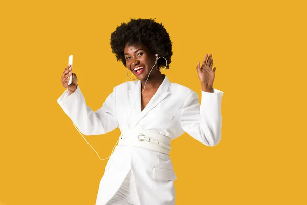 Smiling african american girl listening music while holding smartphone and gesturing isolated on orange — Stock Photo