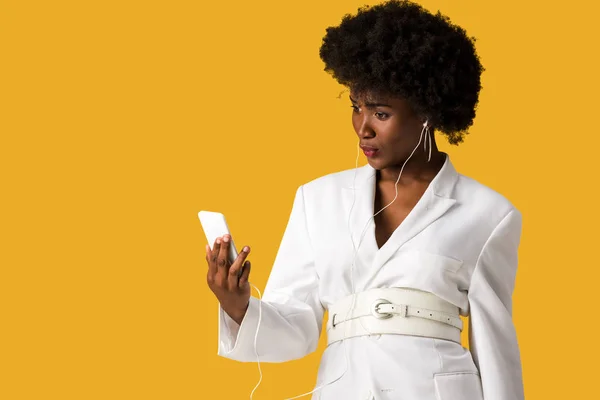 Удивленный африканский американец женщина держит смартфон и слушать музыку изолированы на оранжевый — стоковое фото