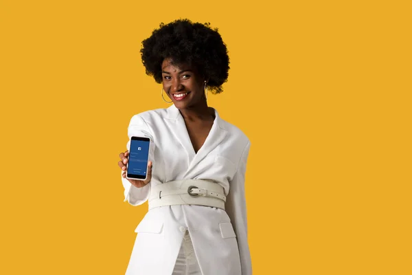 KYIV, UCRANIA - 9 de agosto de 2019: niña afroamericana feliz sosteniendo teléfono inteligente con aplicación de facebook en la pantalla aislada en naranja - foto de stock