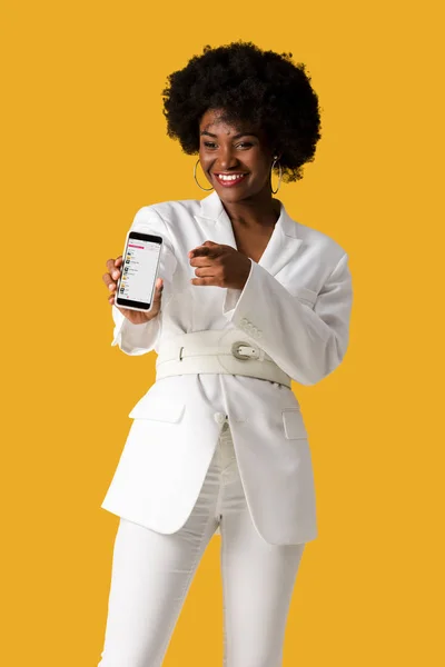 KYIV, UKRAINE - 9 AOÛT 2019 : heureuse fille afro-américaine tenant un smartphone avec application de musique aux pommes à l'écran tout en pointant du doigt isolé sur orange — Photo de stock