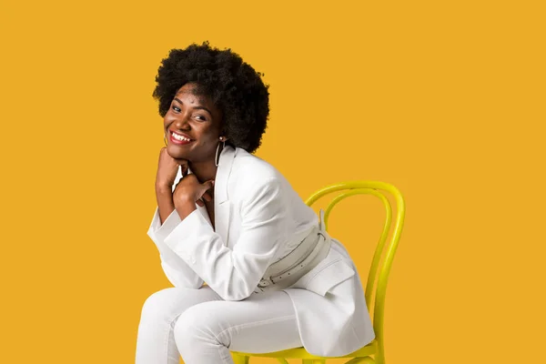 Happy african american girl sitting on yellow chair isolated on orange — Stock Photo