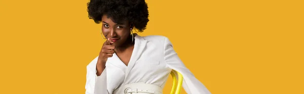 Panoramic shot of african american woman sitting on yellow chair and pointing with finger isolated on orange — Stock Photo