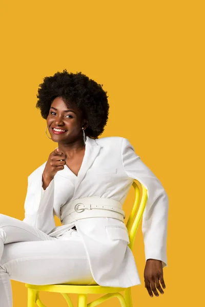 Heureuse femme afro-américaine assise sur chaise jaune et pointant du doigt isolé sur orange — Stock Photo