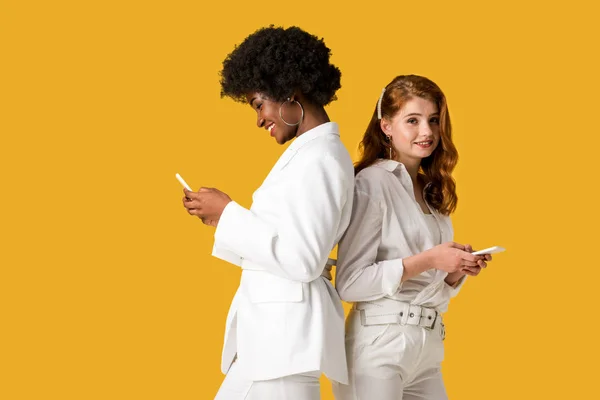 Happy multicultural girls using smartphones while standing isolated on orange — Stock Photo