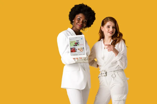KYIV, UCRANIA - 9 de agosto de 2019: alegre pelirroja señalando con el dedo cerca de la mujer afroamericana sosteniendo la tableta digital con la aplicación ebay en la pantalla aislada en naranja - foto de stock
