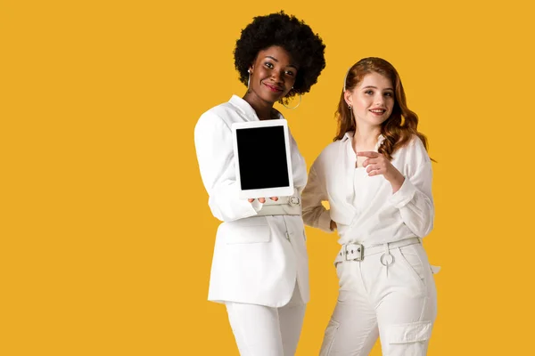 Весела руда дівчина вказує пальцем біля афроамериканки, тримає цифровий планшет з порожнім екраном ізольовано на помаранчевому — стокове фото