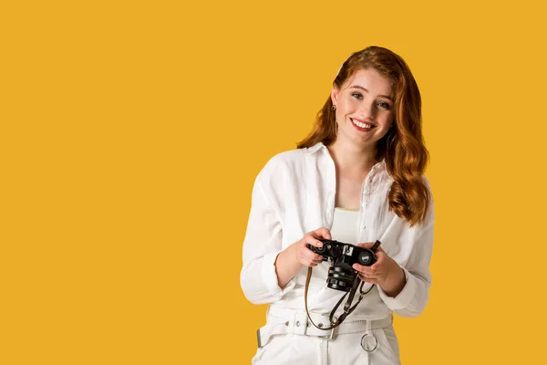 Sonriente pelirroja chica sosteniendo cámara digital aislado en naranja - foto de stock