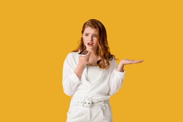 Dissatisfied redhead girl holding smartphone and gesturing isolated on orange — Stock Photo