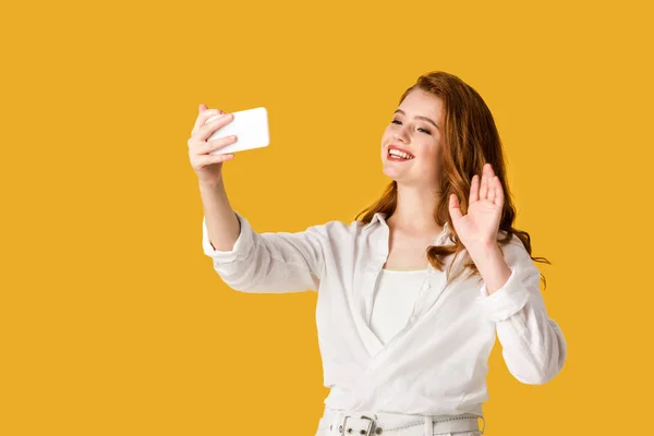 Feliz pelirroja chica tomando selfie y agitando mano aislado en naranja - foto de stock