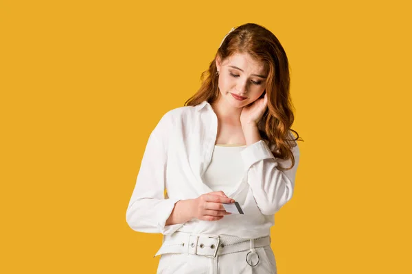 Upset redhead woman looking at credit card isolated on orange — Stock Photo