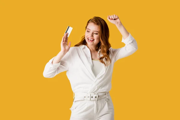 Cheerful redhead girl gesturing while holding credit card isolated on orange — Stock Photo