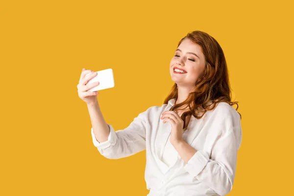 Bonita pelirroja chica tomando selfie y sonriendo aislado en naranja - foto de stock