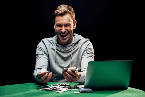 Homem feliz segurando fichas de poker perto de laptop na mesa de poker isolado em preto — Fotografia de Stock