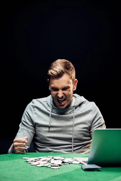 Homme en colère avec les poings serrés près de l'ordinateur portable sur la table de poker isolé sur noir — Photo de stock