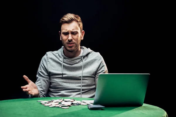 Засмучений чоловік жестикулює біля ноутбука і покерних чіпів ізольовані на чорному — стокове фото