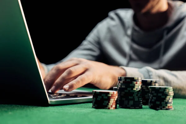 Vista cortada de homem digitando no laptop perto de fichas de poker isolado no preto — Fotografia de Stock
