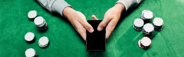 Plano panorámico del hombre sosteniendo teléfono inteligente con pantalla en blanco cerca de fichas de póquer - foto de stock