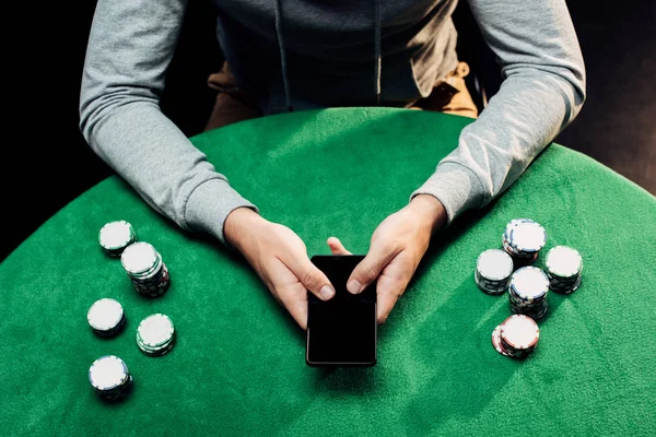 Vista recortada del hombre sosteniendo teléfono inteligente con pantalla en blanco cerca de fichas de póquer en negro - foto de stock