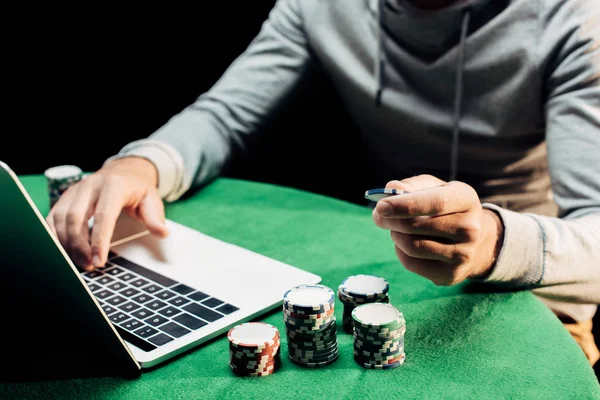 Vista recortada del hombre sosteniendo moneda de póquer y escribiendo en el ordenador portátil aislado en negro - foto de stock