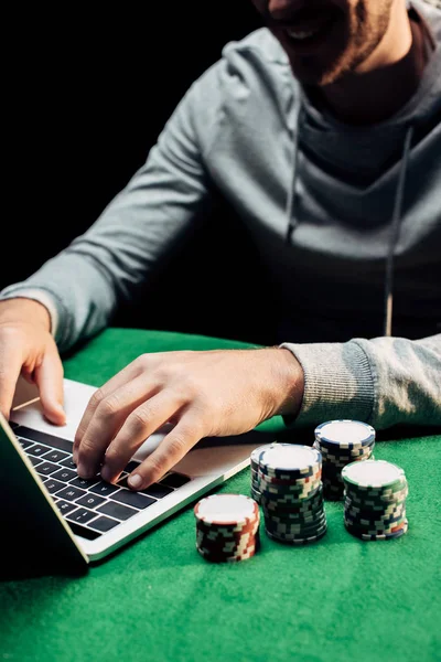 Vista cortada de homem feliz digitando no laptop perto de fichas de poker isolado no preto — Fotografia de Stock
