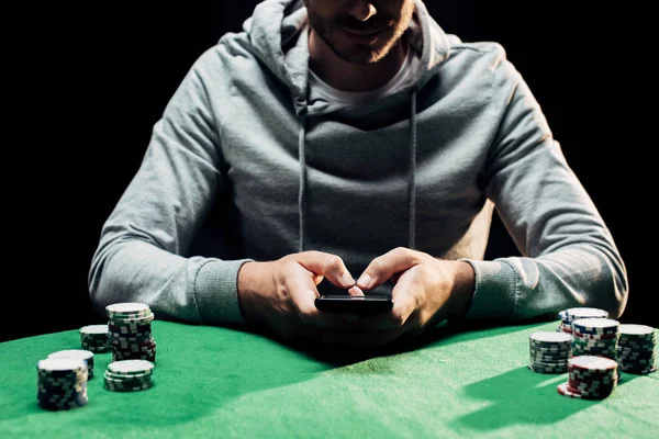 Vista ritagliata di uomo in possesso di smartphone con schermo vuoto vicino a chip di poker isolati su nero — Foto stock
