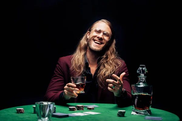 Selektiver Fokus eines glücklichen bärtigen Mannes mit Brille, der ein Glas mit Whiskey in der Nähe von Spielkarten hält, die auf schwarz isoliert sind — Stockfoto