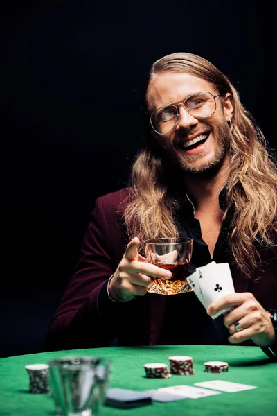 KYIV, UCRANIA - 20 de agosto de 2019: enfoque selectivo del hombre alegre en gafas cerca de jugar a las cartas en la mesa de póquer aislado en negro - foto de stock