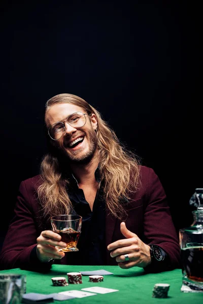 Вибірковий фокус бородатого чоловіка в окулярах, що тримає склянку біля гральних карт на покерному столі ізольовані на чорному — стокове фото