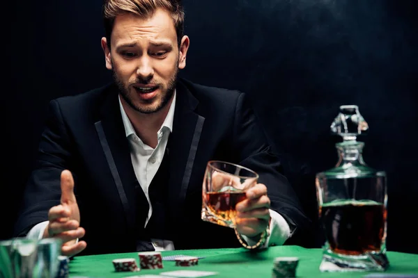 Selective focus of upset man holding glass and gesturing on black with smoke — Stock Photo