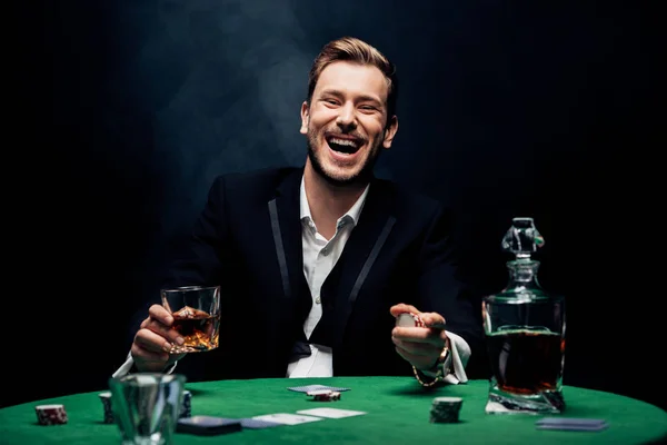 Selective focus of cheerful man holding glass with alcohol on black with smoke — Stock Photo