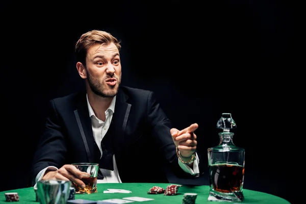 Selective focus of angry man pointing with finger near alcohol and playing cards isolated on black — Stock Photo